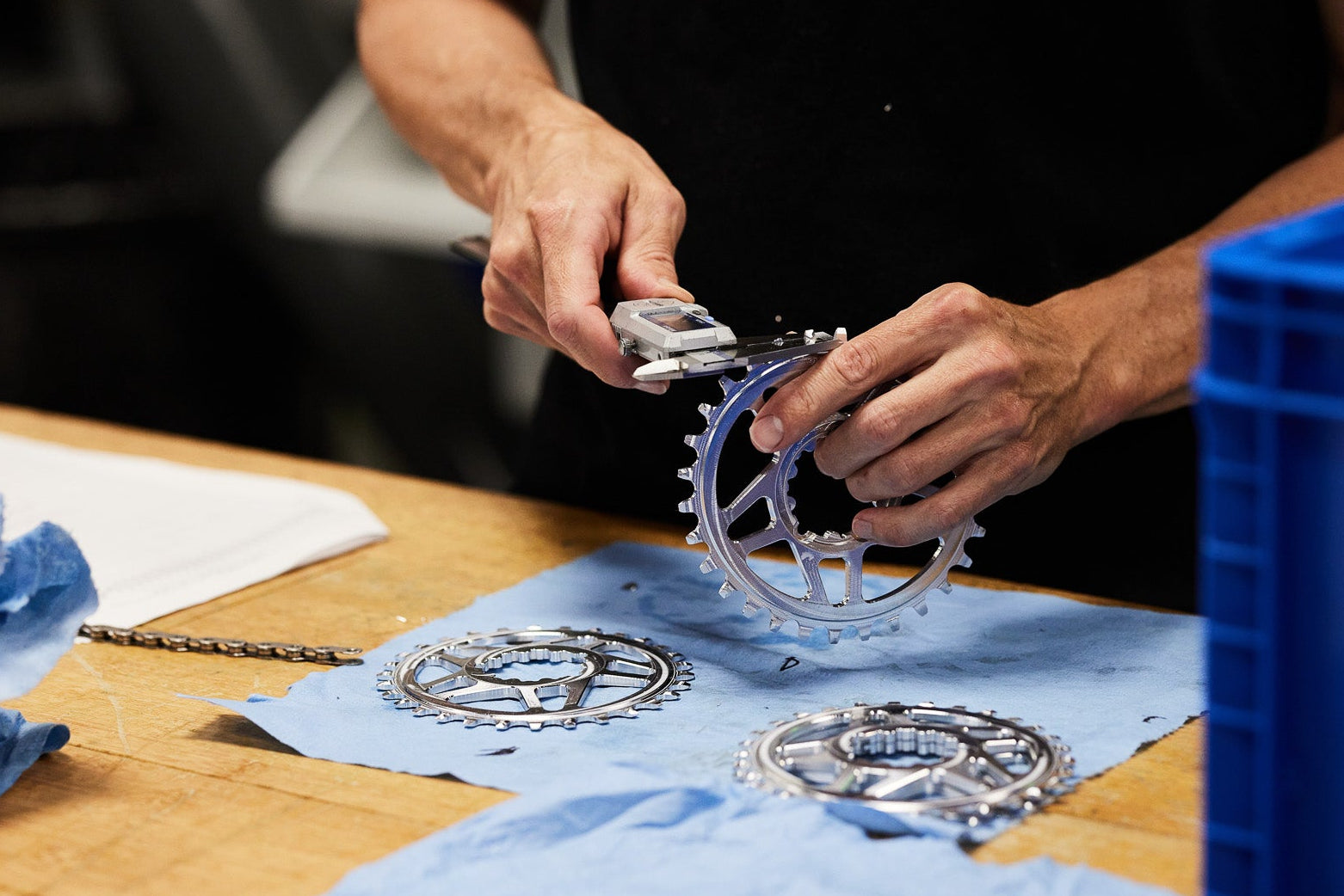 A person in a black shirt holds a silver bicycle chainring and chain on a workbench, with two more chainrings and blue cloth pieces nearby, alongside a large blue plastic bin.
