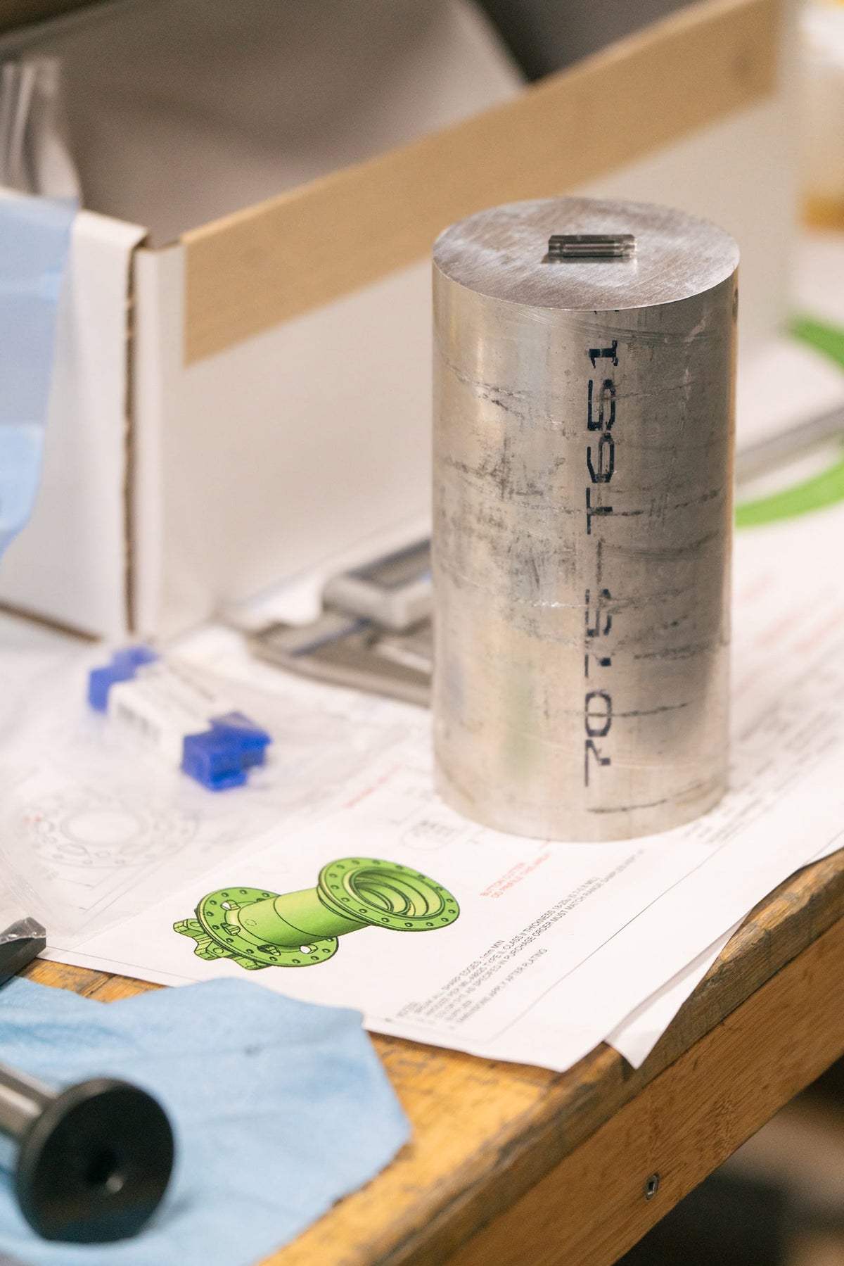 A cylindrical metal part with the numbers '7075-T651' stamped vertically on its side, standing on a workbench next to technical drawings of a Wolf Tooth hub.